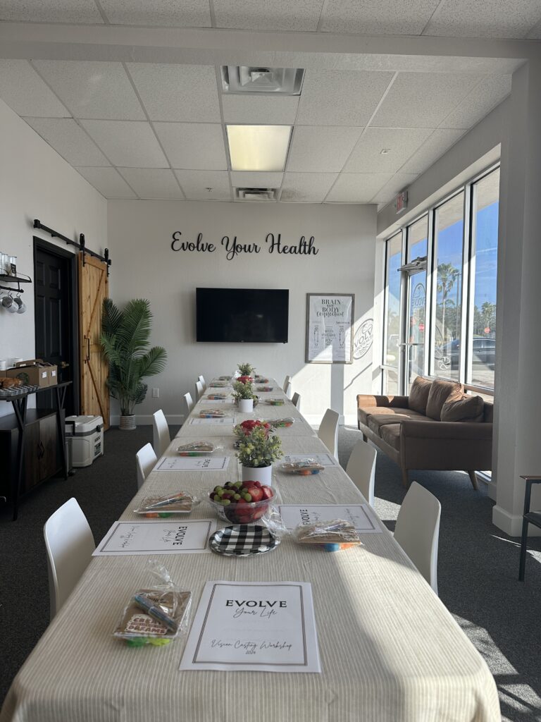 Long table set up with a linen table cloth, spring flowers, fruit plates, and a paper workbook for each participant at their corresponding seats. all of this is set up in the front office of evolve chiropractic